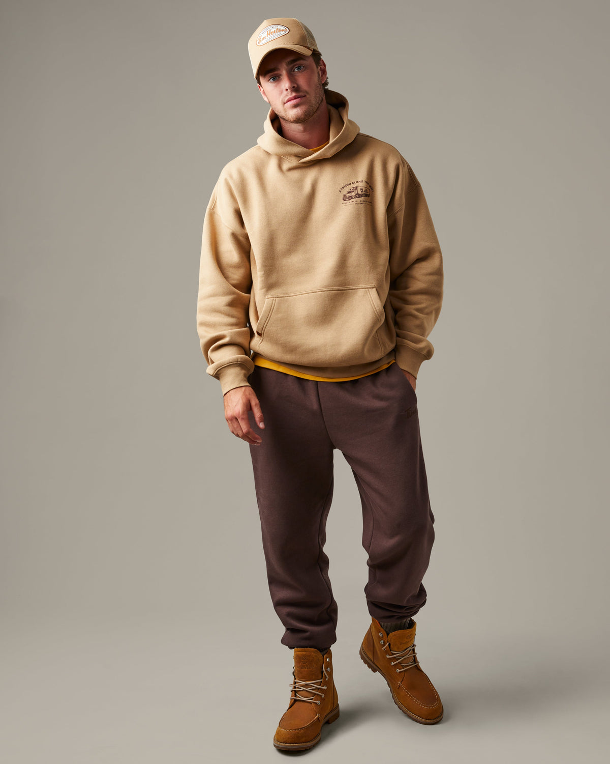 Beige Tim Hortons pullover hoodie with a kangaroo pocket and small A Friend Along the Way delivery truck graphic on the chest. | Chandail à capuchon Tim Hortons beige avec poche kangourou et petite illustration de camion de livraison « Un ami sur la route » sur la poitrine.
